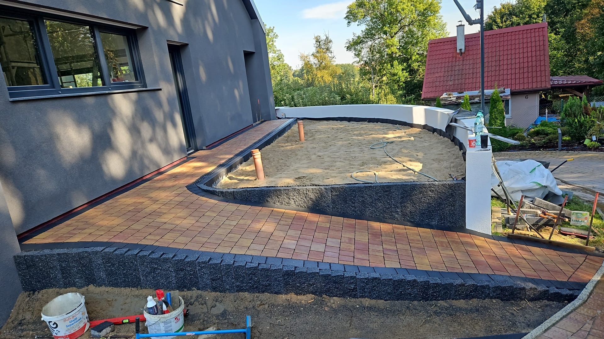 Ułożona kostka brukowa w odcieniach czerwieni i brązu, obramowana czarnym obrzeżem, prowadząca do obszaru wypełnionego piaskiem z wystającymi rurami PCV, widoczne narzędzia i materiały budowlane...