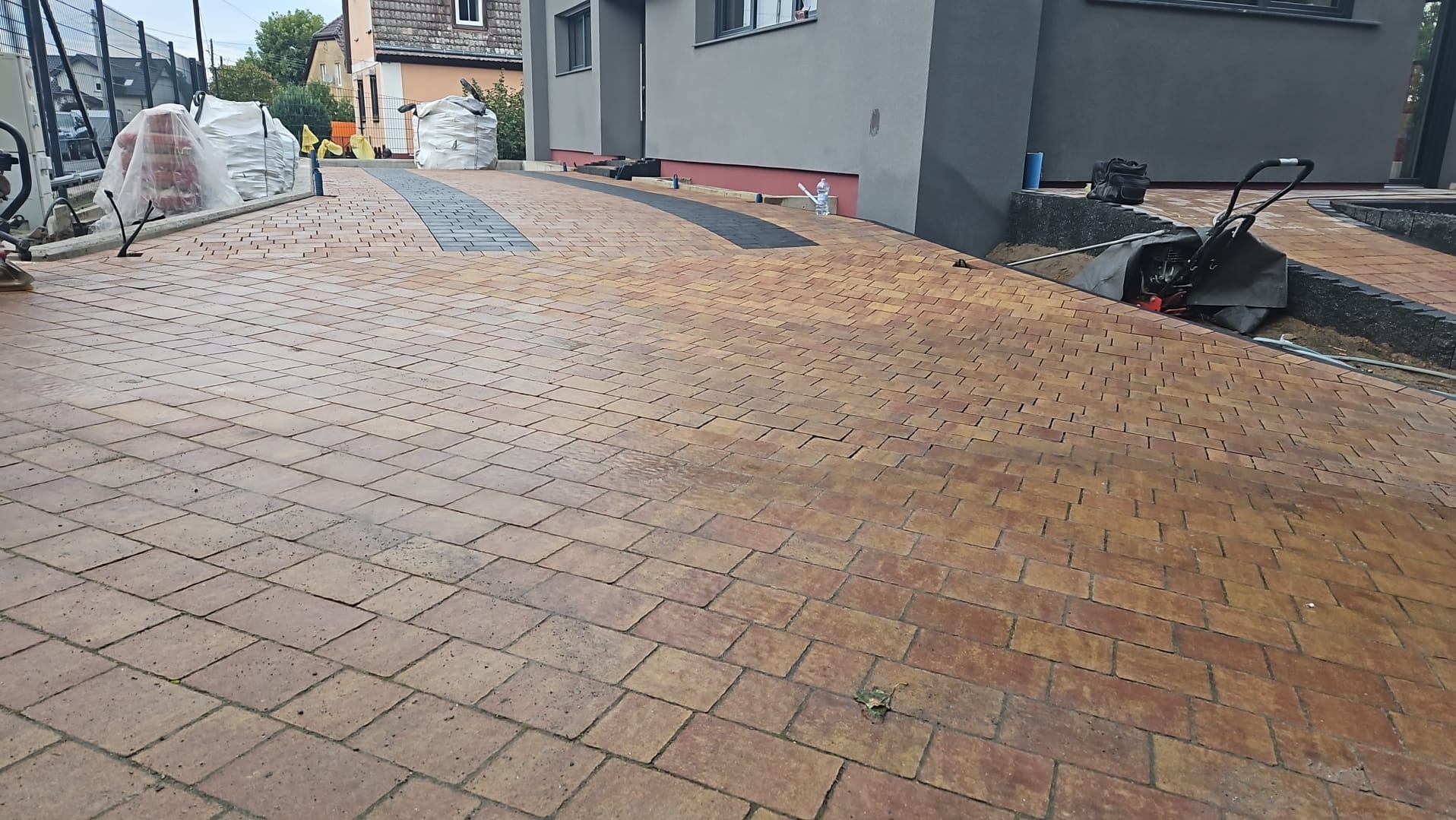 Nowo ułożona, brązowo-pomarańczowa kostka brukowa na podjeździe do nowoczesnego domu z szarą elewacją, z widocznymi workami z materiałem budowlanym i częściowo ukrytą maszyną do zagęszczania gruntu.