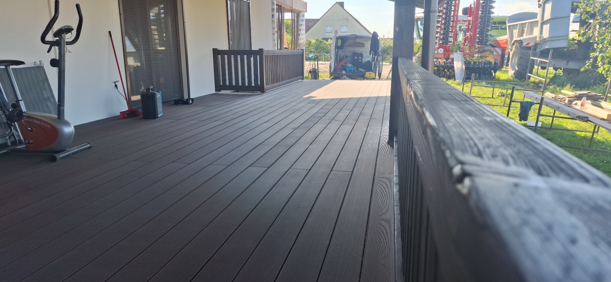 Nowoczesny taras z kompozytowej deski w odcieniach brązu, z ciemną balustradą i widokiem na podwórze z zaparkowanym busem i sprzętem rolniczym, w tle jasny budynek.