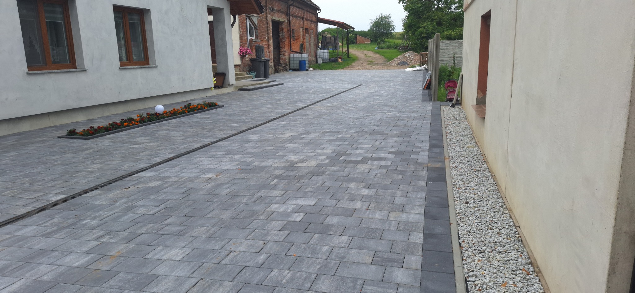 Ułożona szara kostka brukowa na podjeździe z liniowym odpływem, obok rabata kwiatowa i pas z białym grysem przy ścianie budynku.