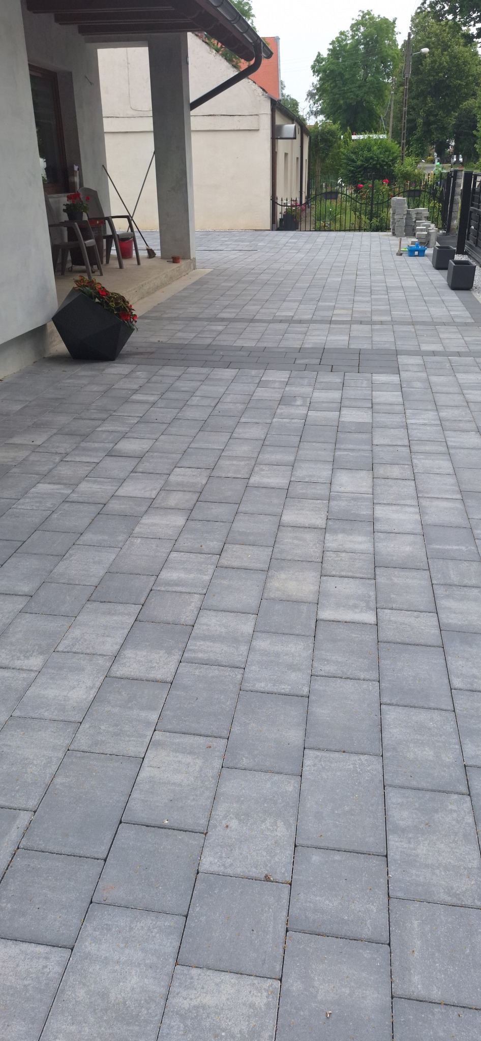 Ułożona szara kostka brukowa na podjeździe przed domem z widocznymi donicami z kwiatami i częścią ogrodu w tle.