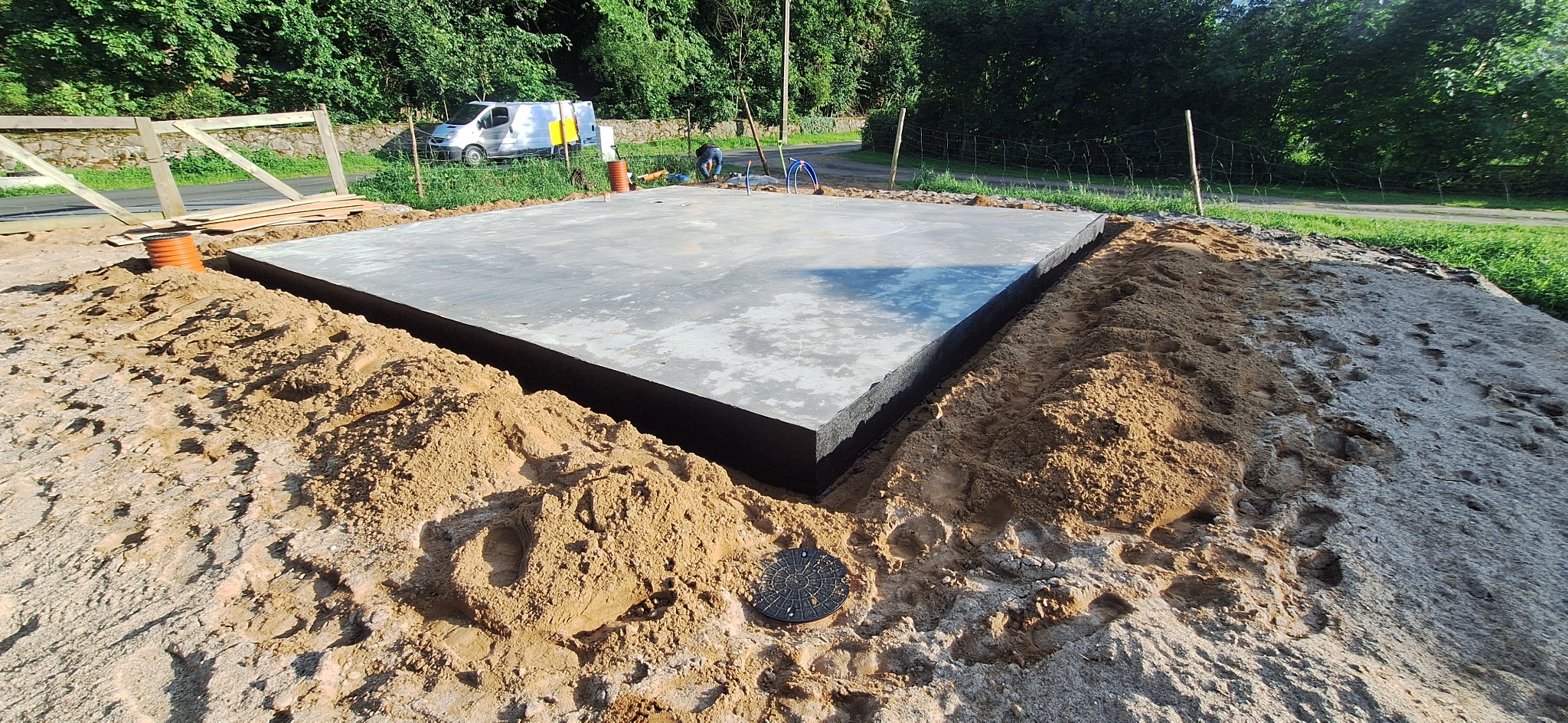 Wylany fundament z izolacją pionową, otoczony piaskiem. W tle widoczny bus i zieleń. Na pierwszym planie studzienka rewizyjna.