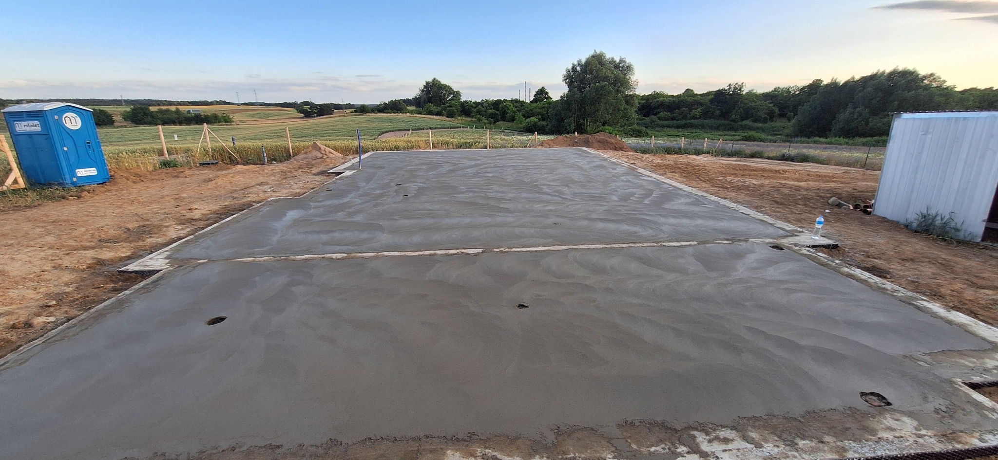 Wylany fundament pod budynek z widocznymi otworami. W tle pola uprawne i las. Na skraju kadru TOI TOI i budynek gospodarczy. Niebo lekko zachmurzone.