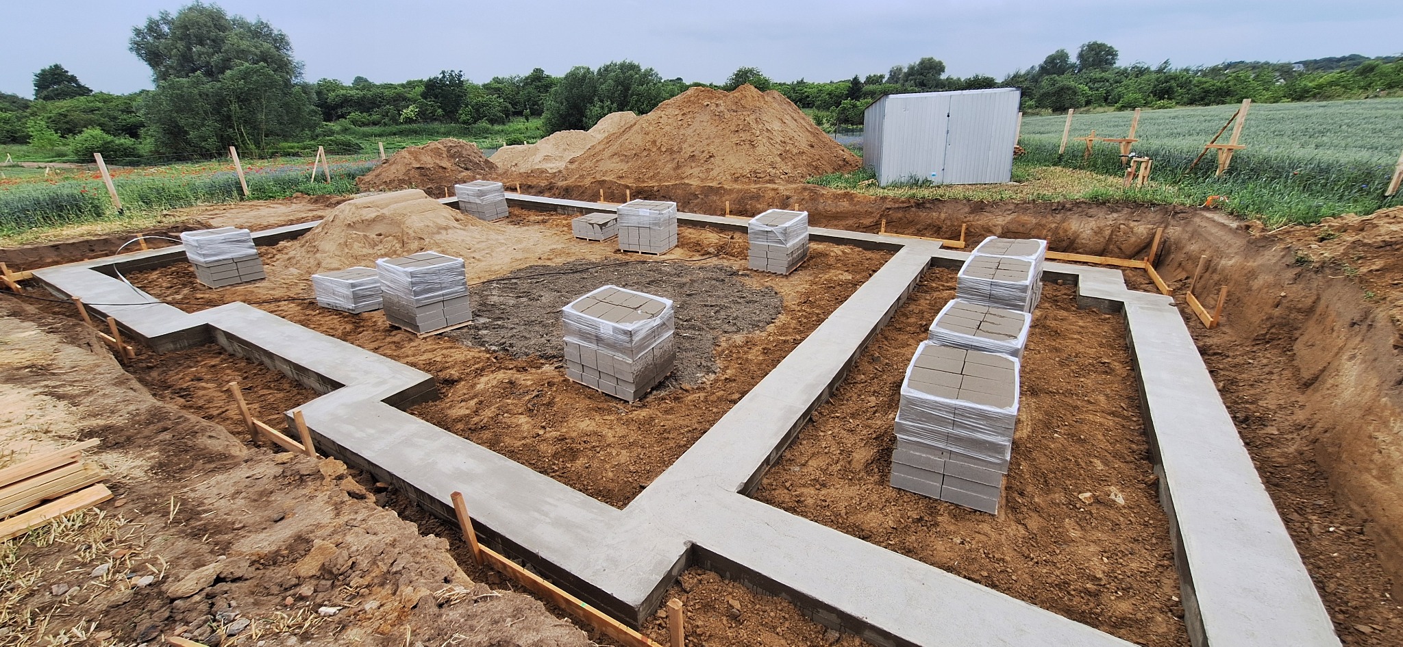 Zalane fundamenty pod budynek z ułożonymi bloczkami w folii, widok z góry na wykop z drewnianym szalunkiem. W tle hałda ziemi i blaszany garaż.
