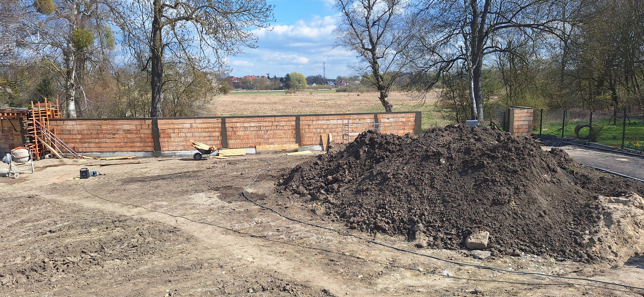 Wznoszenie muru z cegły klinkierowej na fundamencie. Widoczna sterta ziemi, mieszarka i narzędzia budowlane. Tło stanowi krajobraz z drzewami i budynkami.