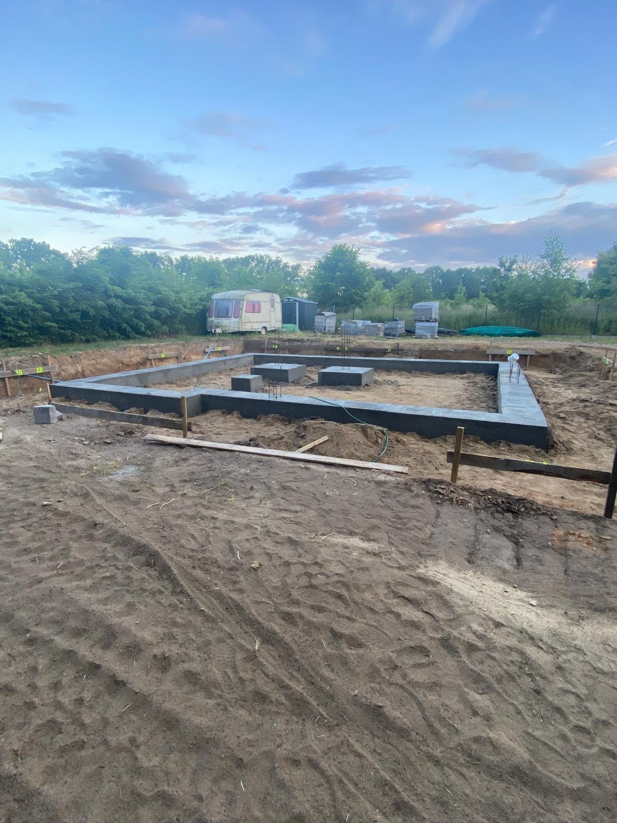Wylane fundamenty pod budowę domu z widocznymi zbrojeniami i deskowaniem. W tle przyczepa kempingowa i drzewa.