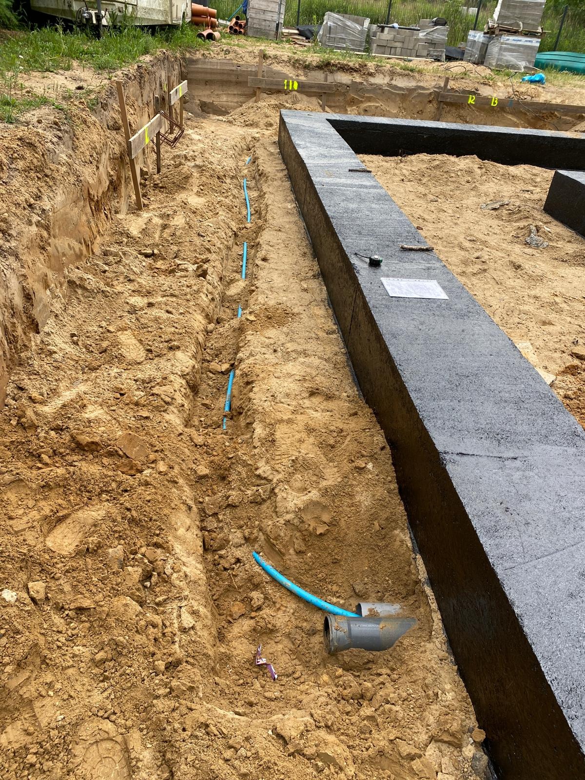 Wykop pod fundamenty z izolacją i niebieskimi rurami instalacji wodnej w ziemi. Widoczne narzędzia pomiarowe i dokumentacja.