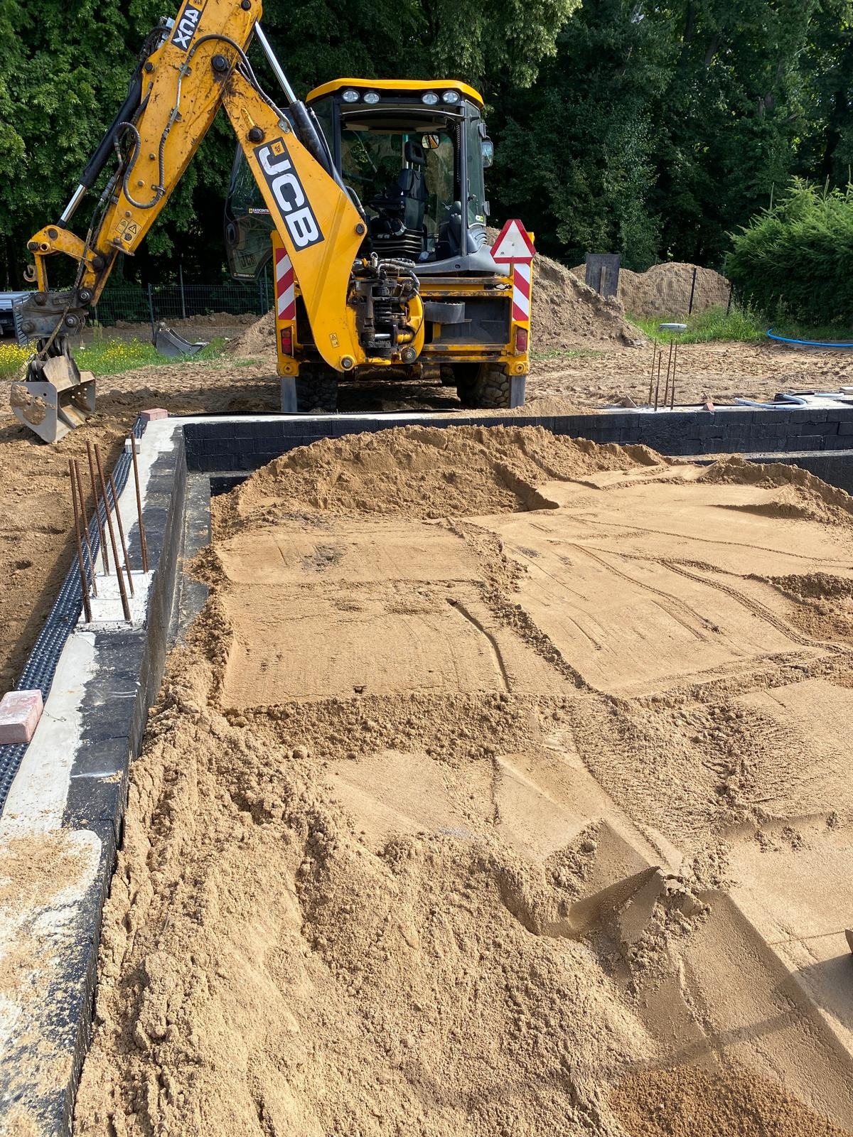 Żółta koparka JCB zasypuje piaskiem fundamenty domu z wystającymi prętami zbrojeniowymi; widoczne ślady pracy maszyny.
