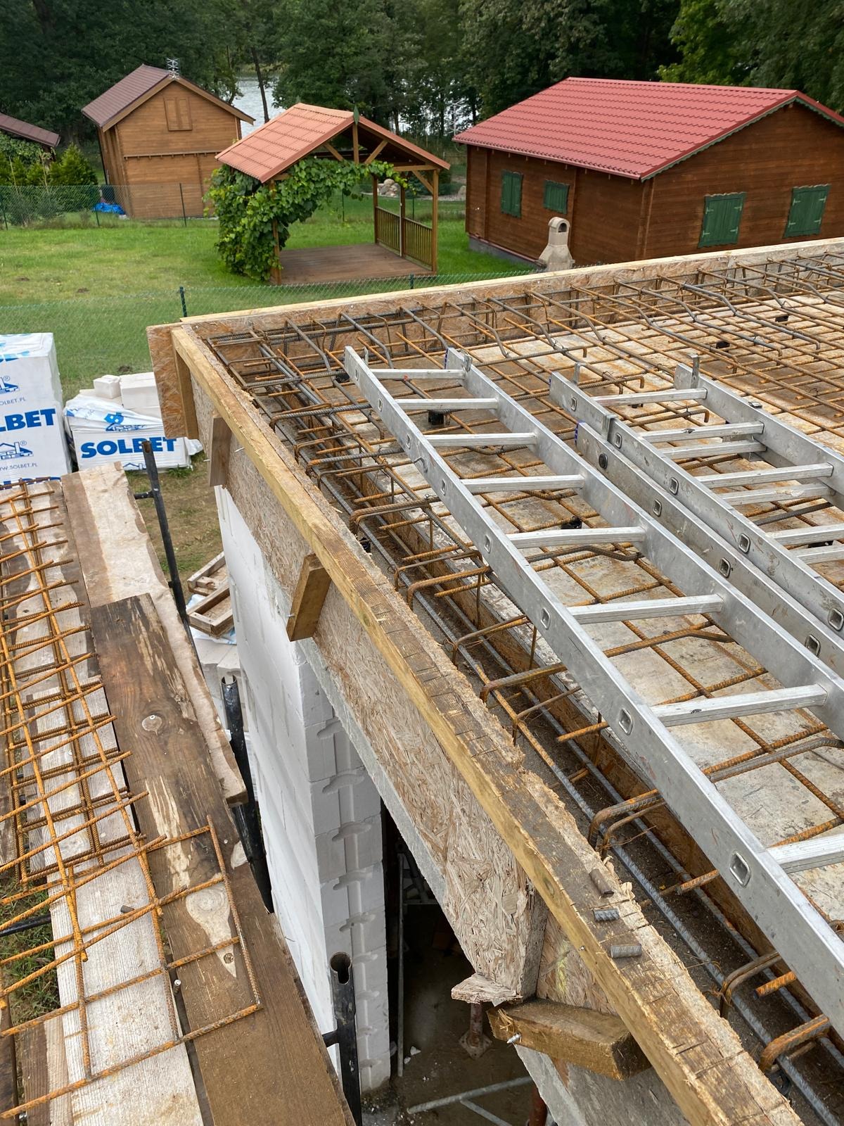 Zbrojenie stropu domu jednorodzinnego z białych bloczków, widoczne drabiny i materiały budowlane. W tle domki letniskowe i drzewa.