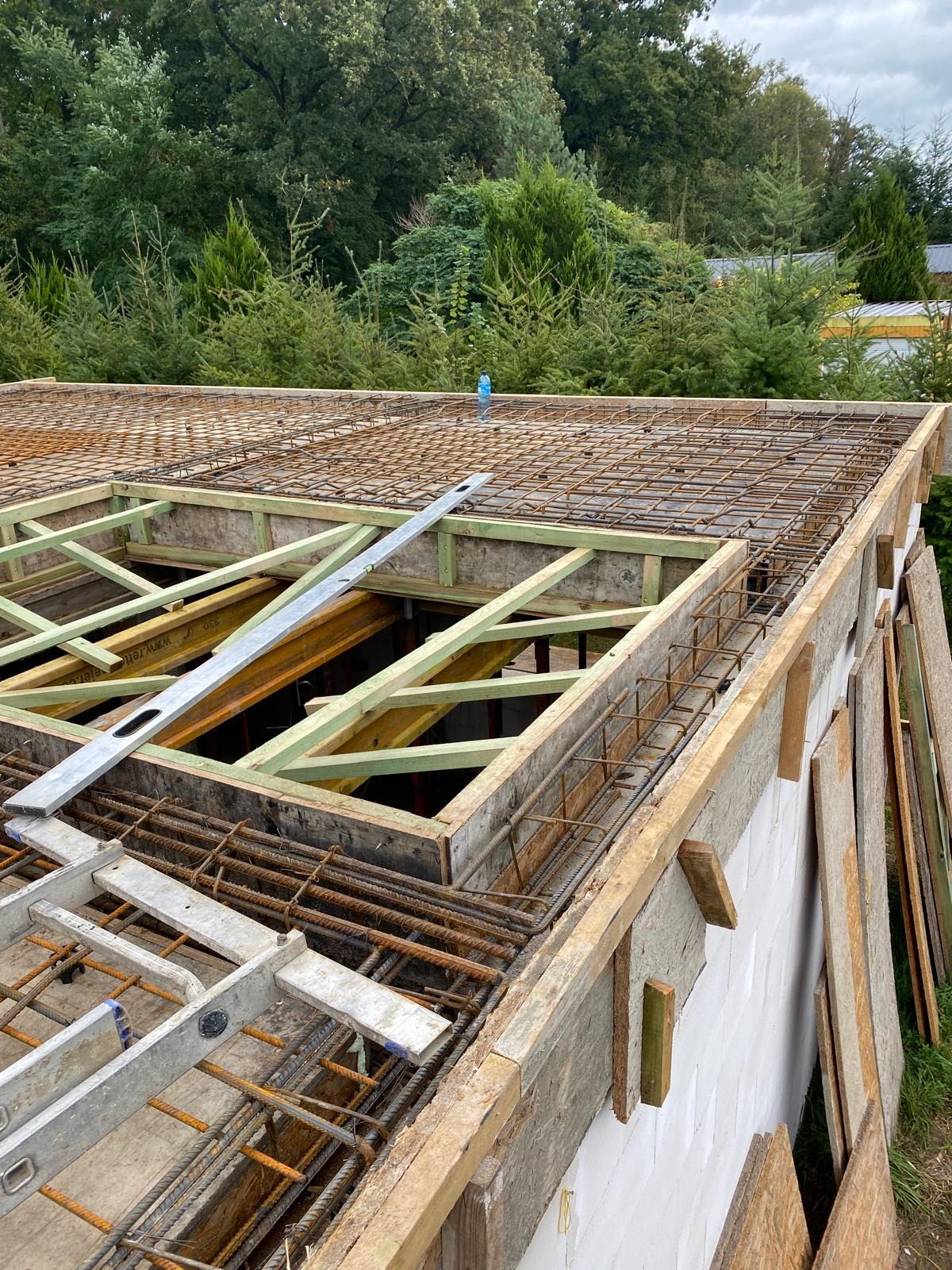 Zbrojenie stropu budynku w trakcie budowy, widoczne drewniane szalunki, stalowe pręty i poziomica. W tle zieleń drzew.