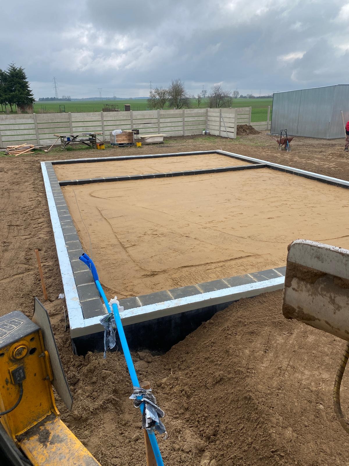 Przygotowany teren pod fundament z częściowo ułożonymi bloczkami, widoczna izolacja i piasek. W tle ogrodzenie panelowe i zabudowania gospodarcze.