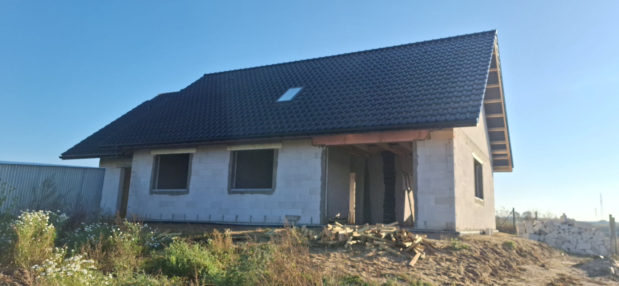 Surowy dom jednorodzinny z ciemnym dachem, widoczne bloczki betonowe i niezadaszony taras. Na pierwszym planie roślinność i ziemia, w tle błękitne niebo.