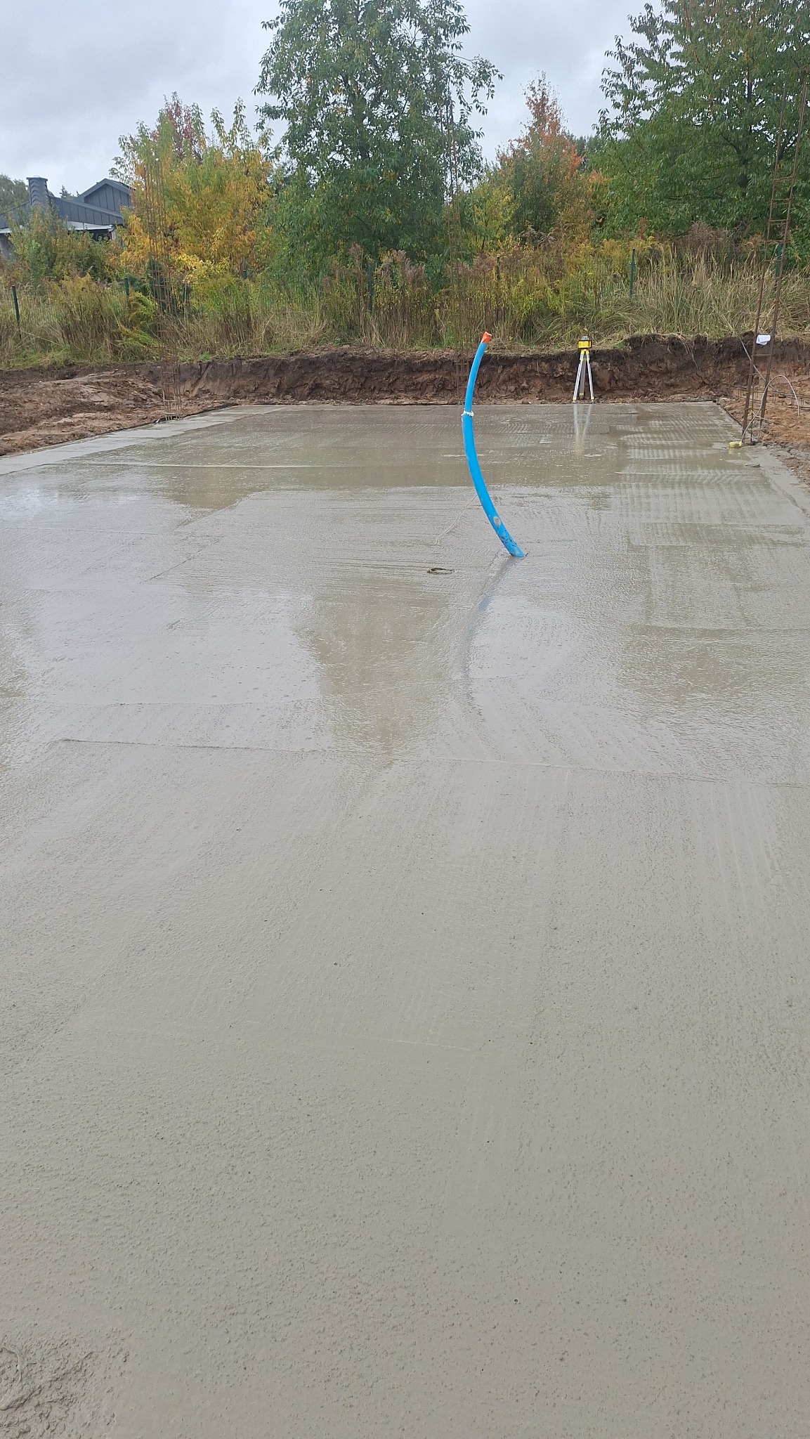 Wylana, gładka płyta fundamentowa z wystającą niebieską rurą instalacyjną. W tle niwelacja terenu i drzewa.