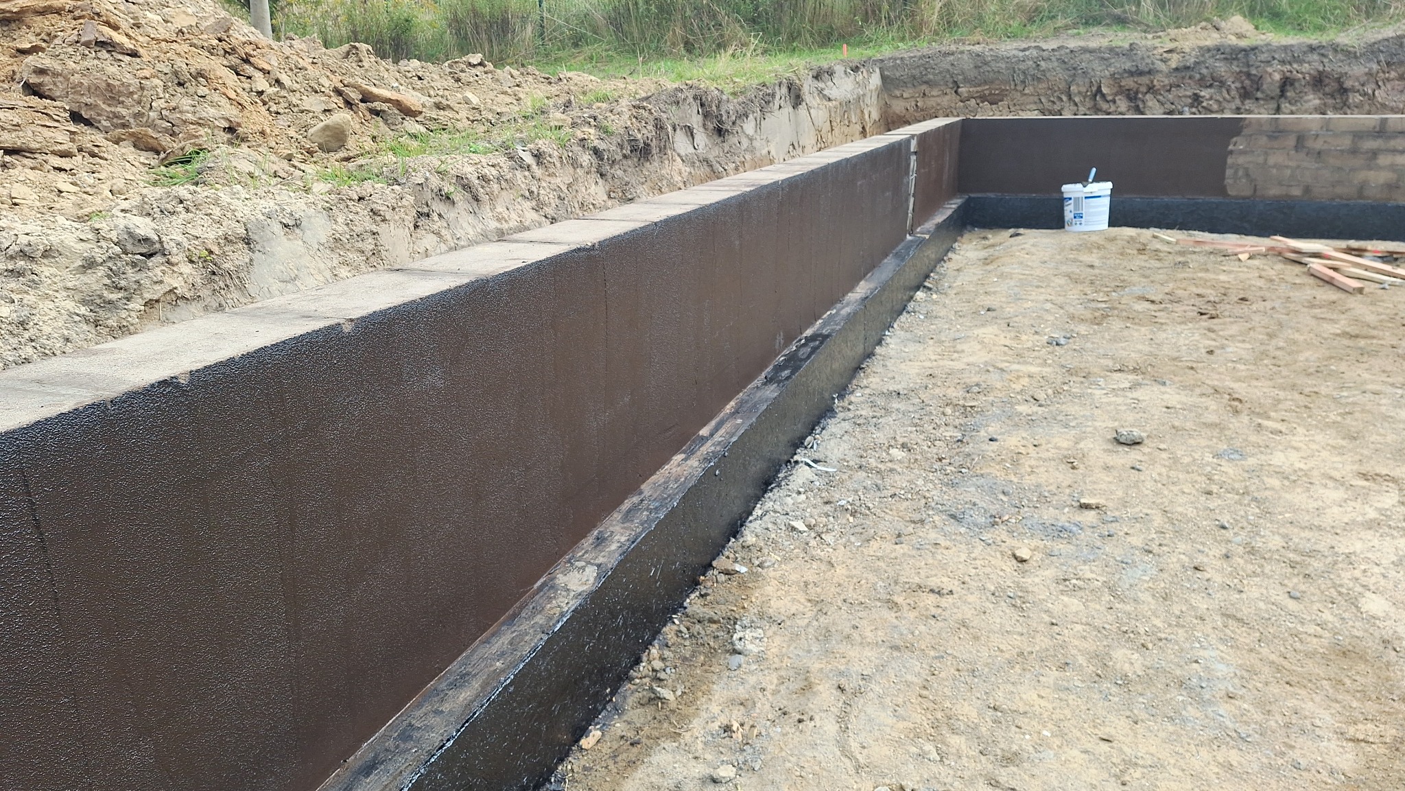 Zabezpieczone hydroizolacją ściany fundamentowe z bloczków betonowych, widoczny grunt i wiadro z narzędziami. Etap prac budowlanych, brązowa powłoka ochronna.