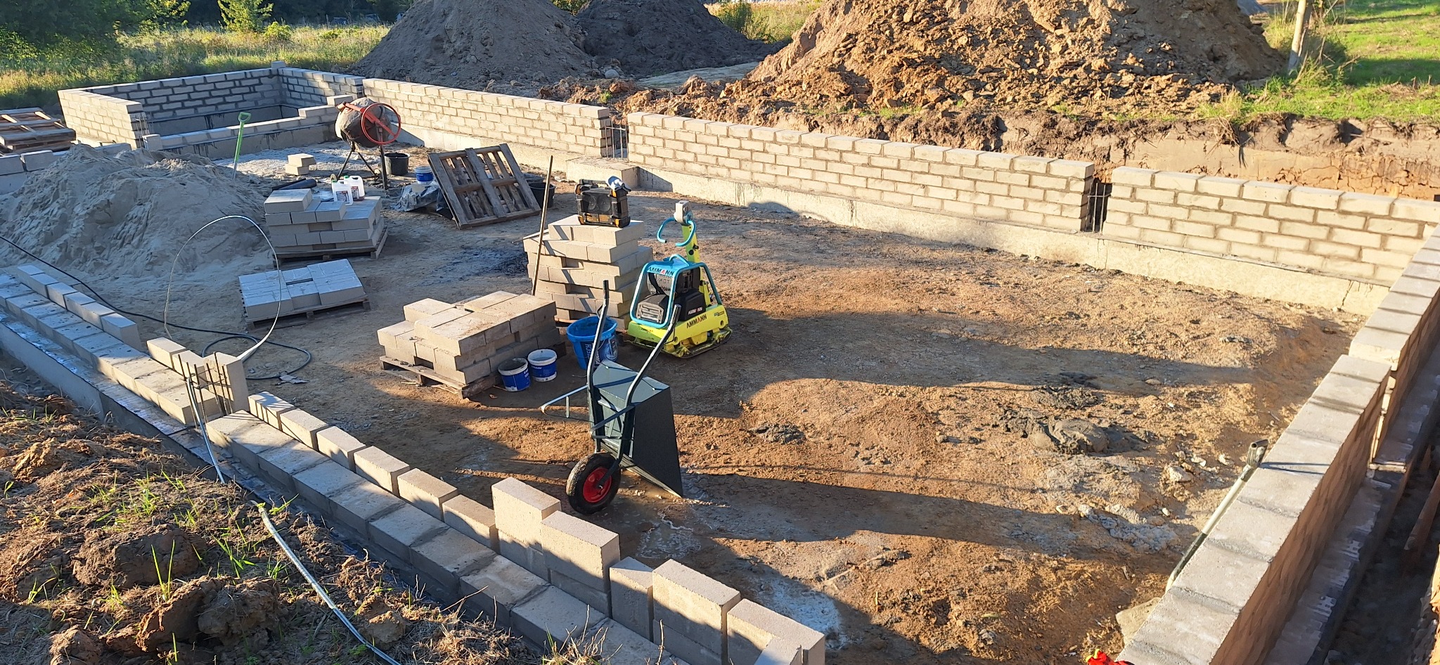 Wykop pod fundamenty z częściowo wymurowanymi ścianami z bloczków betonowych. Na placu budowy widoczny sprzęt budowlany i materiały. Ujęcie z góry, w słoneczny dzień.
