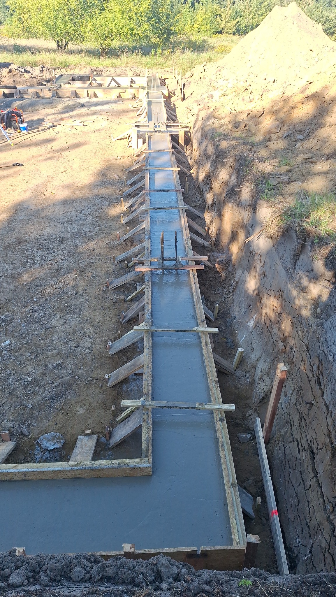 Zalany beton w szalunku drewnianym, widok z góry. W tle wykop ziemny i hałda ziemi. Elementy zbrojenia wystają z betonu. Prace fundamentowe w toku.