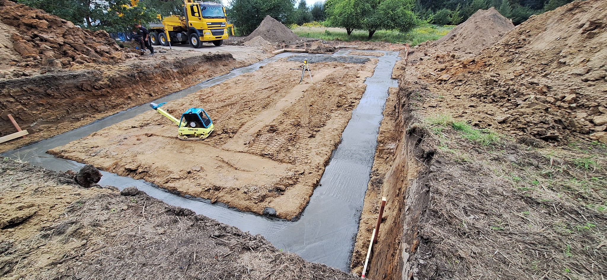 Wykop pod fundamenty z wylaną ławą fundamentową, ubijarką i niwelatorem laserowym na placu budowy. W tle żółty dźwig samochodowy i hałdy ziemi.