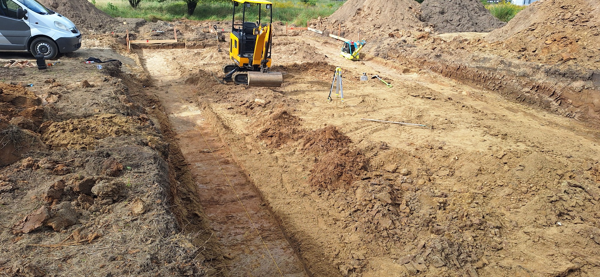 Żółta mini koparka w wykopie pod fundamenty, widoczne narzędzia geodezyjne i hałdy ziemi. Wykop wytyczony sznurkiem, w tle zaparkowany bus.