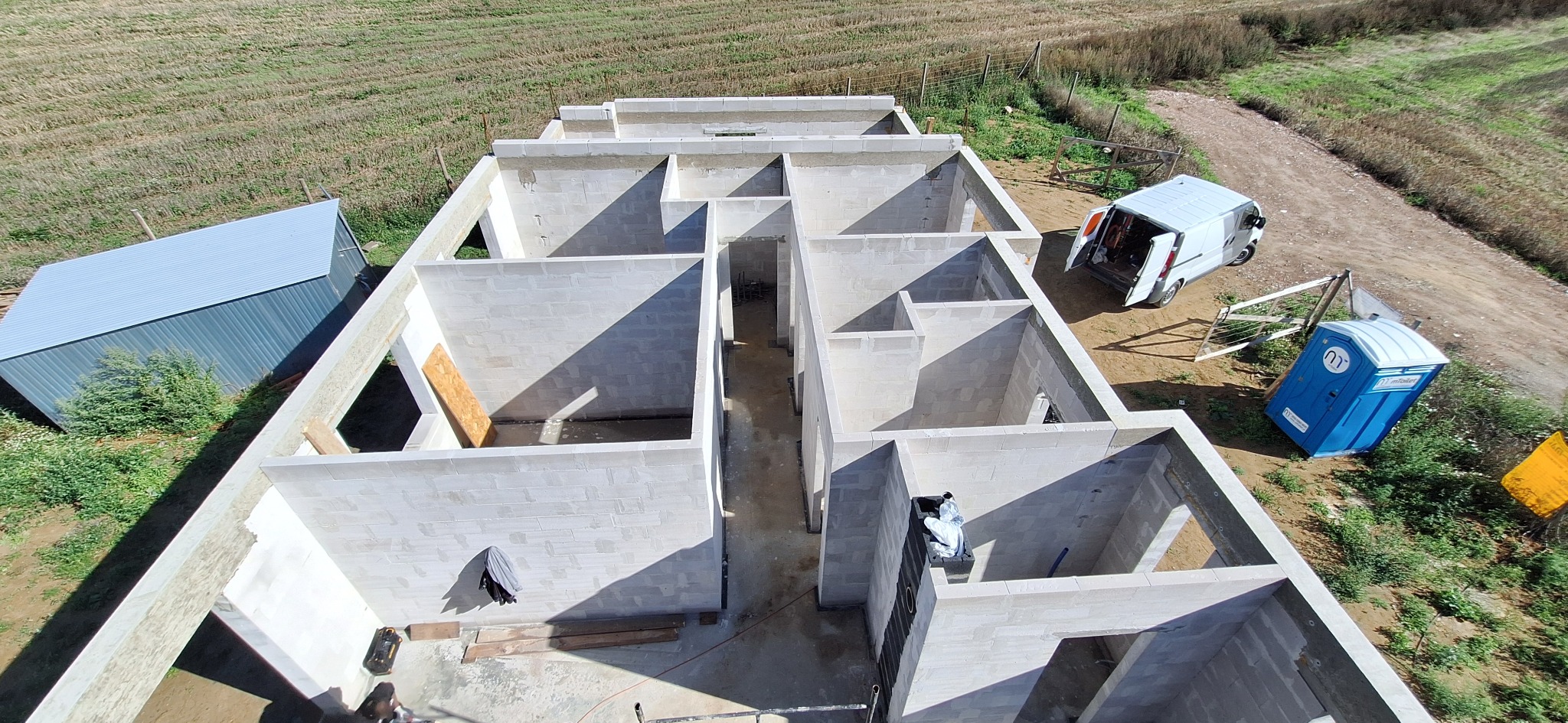 Surowy stan domu z betonu komórkowego z lotu ptaka. Widoczny szary blaszak, biały van z otwartymi drzwiami i niebieska toaleta przenośna na tle pola i drogi.