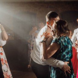 LensStory - Para w objęciach tańczy podczas eventu w Pabianicach, kobieta w okularach i sukience w odcieniach zieleni, mężczyzna w białej koszuli, w tle inni goście i efekty świetlne.