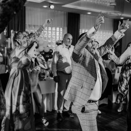 LensStory - Roztańczeni goście w sali balowej podczas uroczystości, mężczyzna w kraciastej marynarce i spodniach na pierwszym planie, uniesione ręce, radosne twarze, czarno-biała fotografia.
