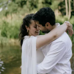 Uśmiechnięta para młoda w objęciach na tle rzeki i zieleni, panna młoda w zwiewnej sukience z delikatnymi złotymi kolczykami.