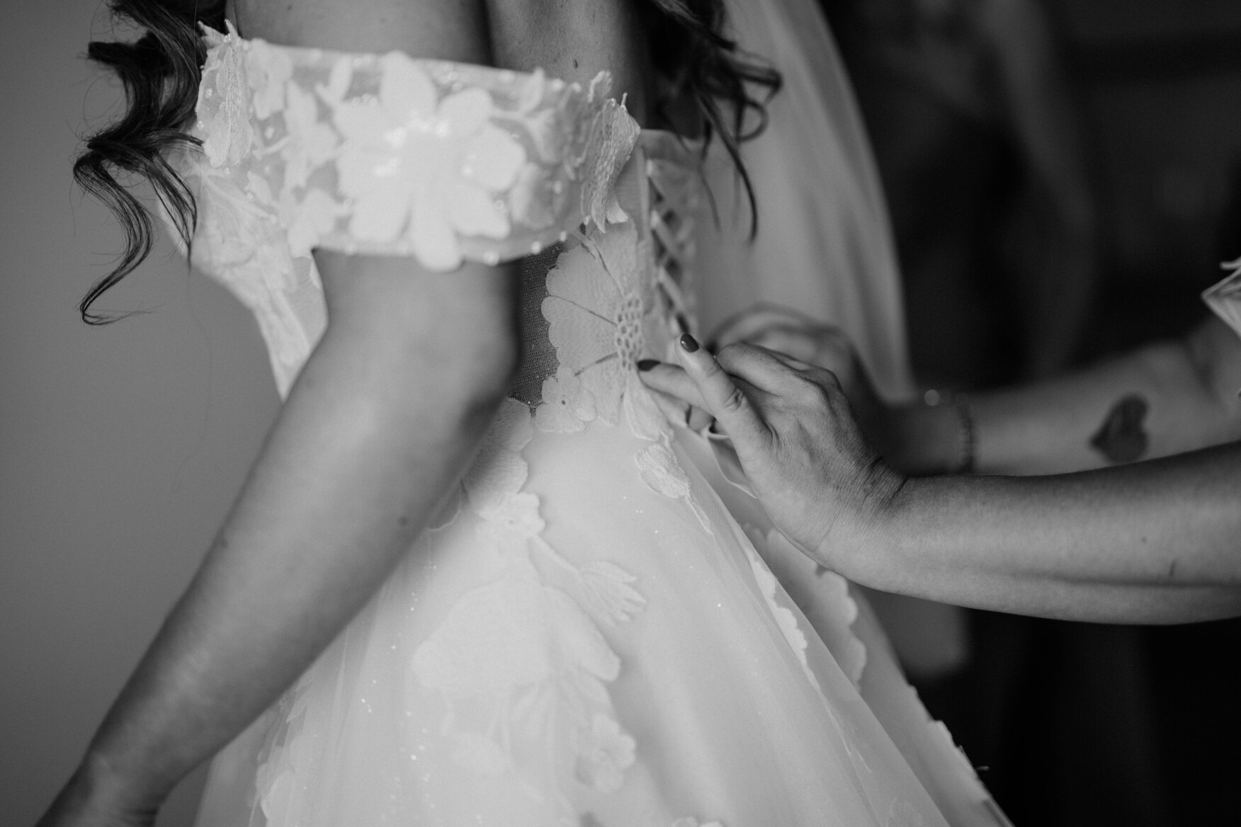 Czarno-białe zbliżenie sukni ślubnej z kwiatowymi aplikacjami, sznurowanej na plecach, z widocznymi dłońmi poprawiającymi sznurowanie i tatuażem serca na ramieniu