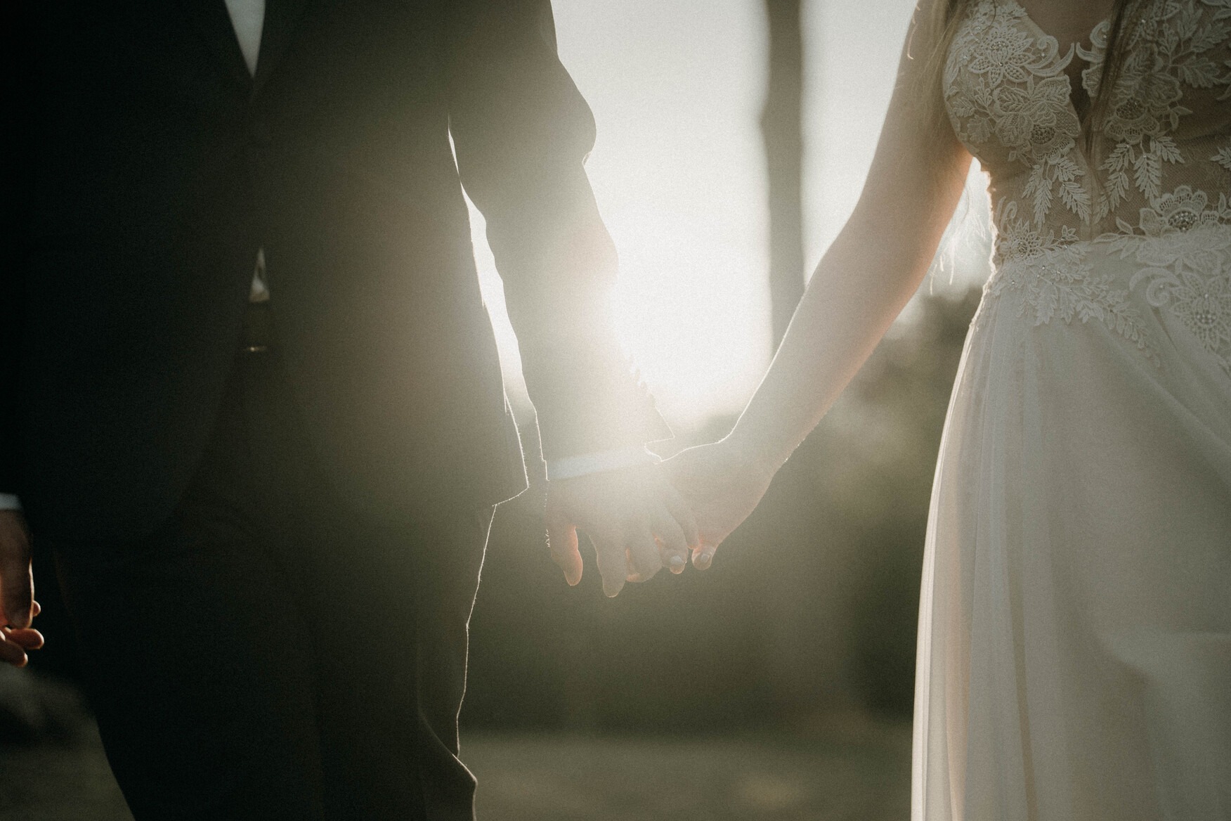 Para trzymająca się za ręce w promieniach zachodzącego słońca, panna młoda w koronkowej sukni, pan młody w ciemnym garniturze, widok od pasa w górę, delikatne rozmycie tła.