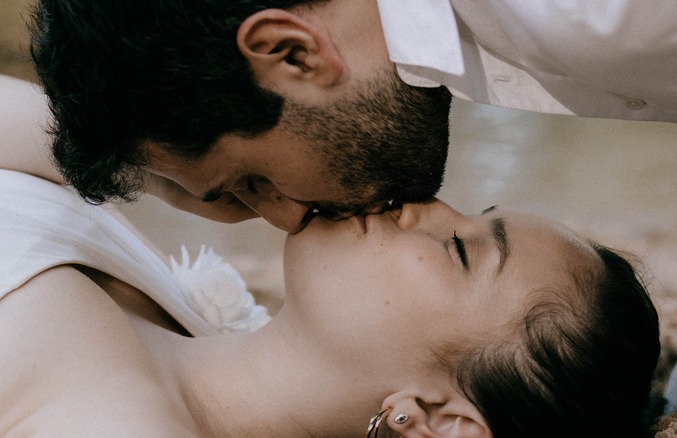 Nowożeńcy w pocałunku, ona z zamkniętymi oczami i złotymi kolczykami kołami, on w białej koszuli, delikatne oświetlenie.