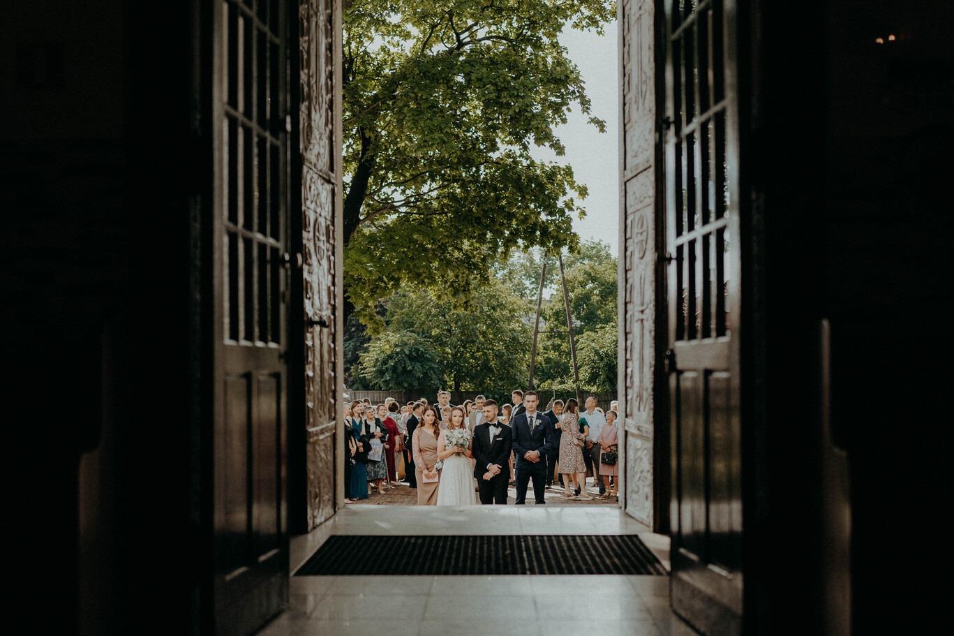 Para młoda wychodzi z kościoła, widziana przez otwarte, zdobione drzwi, z gośćmi weselnymi w tle.