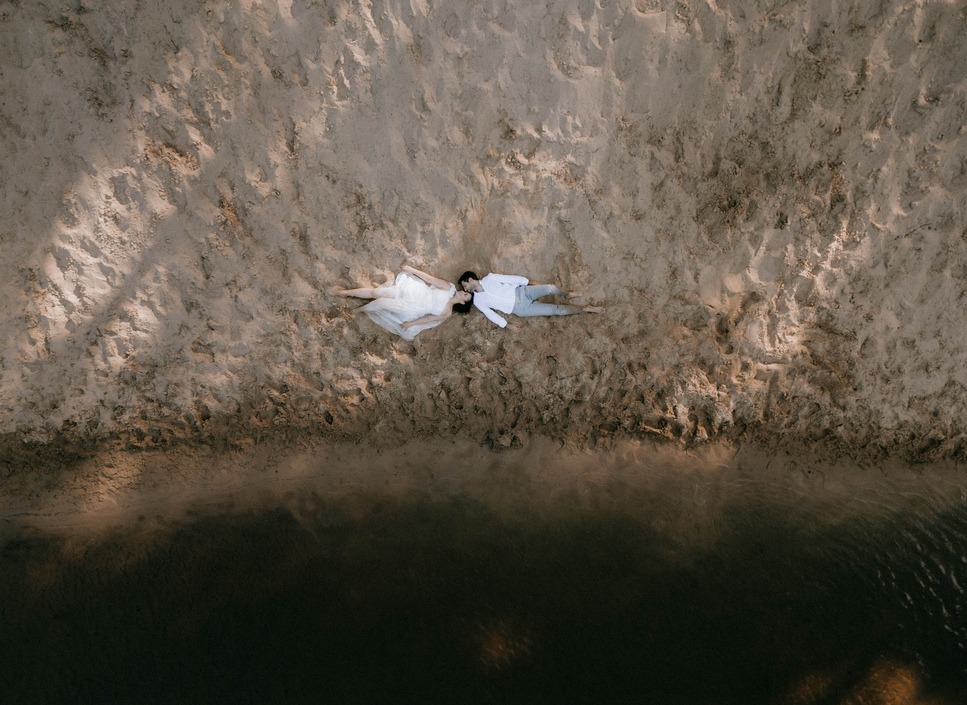 Para młoda leżąca na piasku obok wody, widok z góry, delikatne oświetlenie podkreśla strukturę piasku.