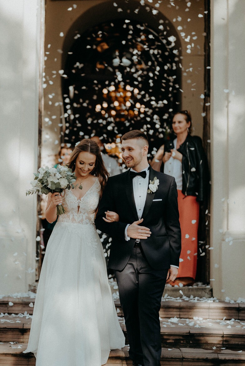 Uśmiechnięta para młoda wychodzi z budynku, obsypywana białymi płatkami konfetti; panna młoda trzyma bukiet białych kwiatów z zielonymi liśćmi, pan młody w czarnym smokingu z muchą.
