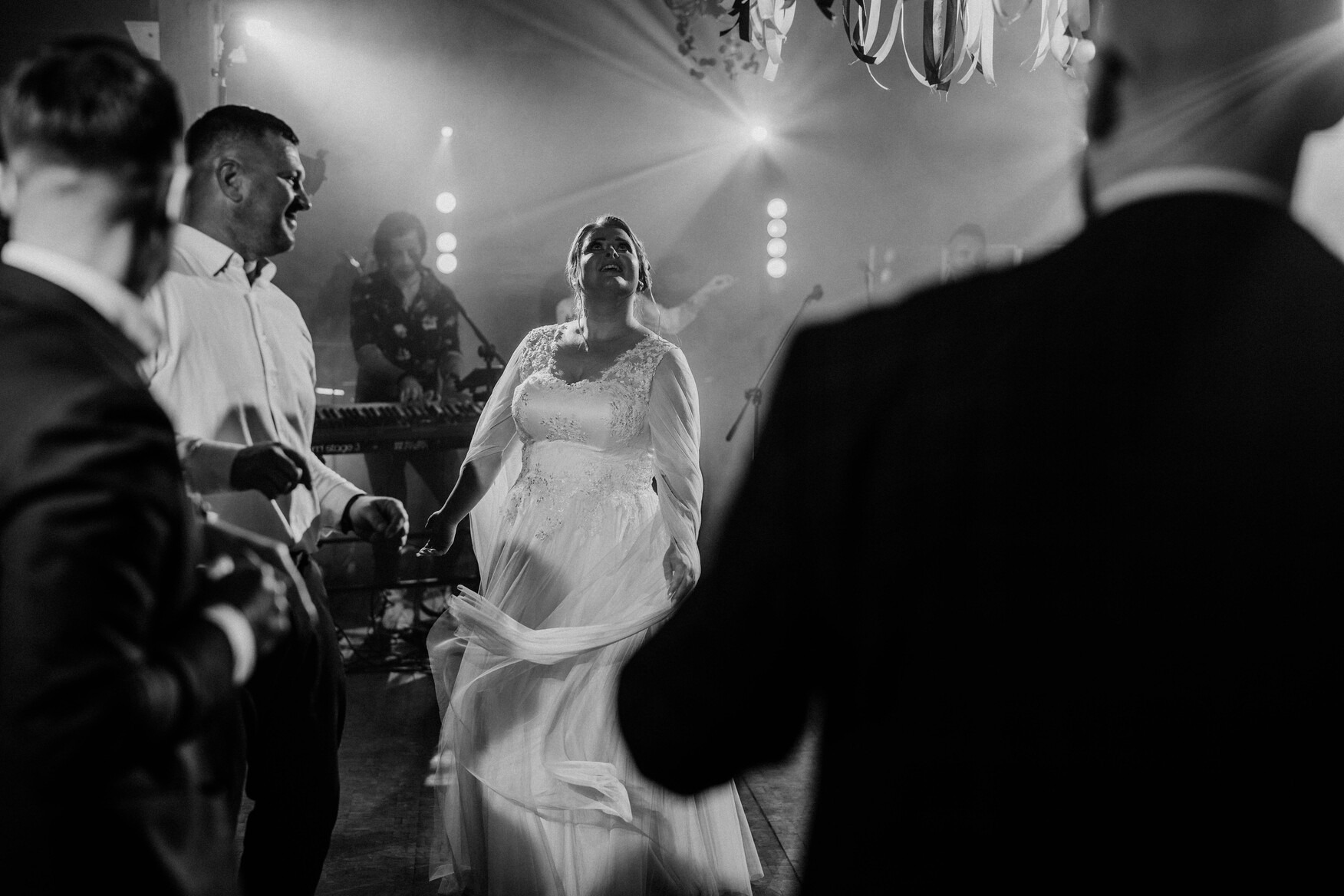 Panna młoda w koronkowej sukni wiruje na parkiecie podczas wesela, w tle zespół muzyczny i dekoracje świetlne.