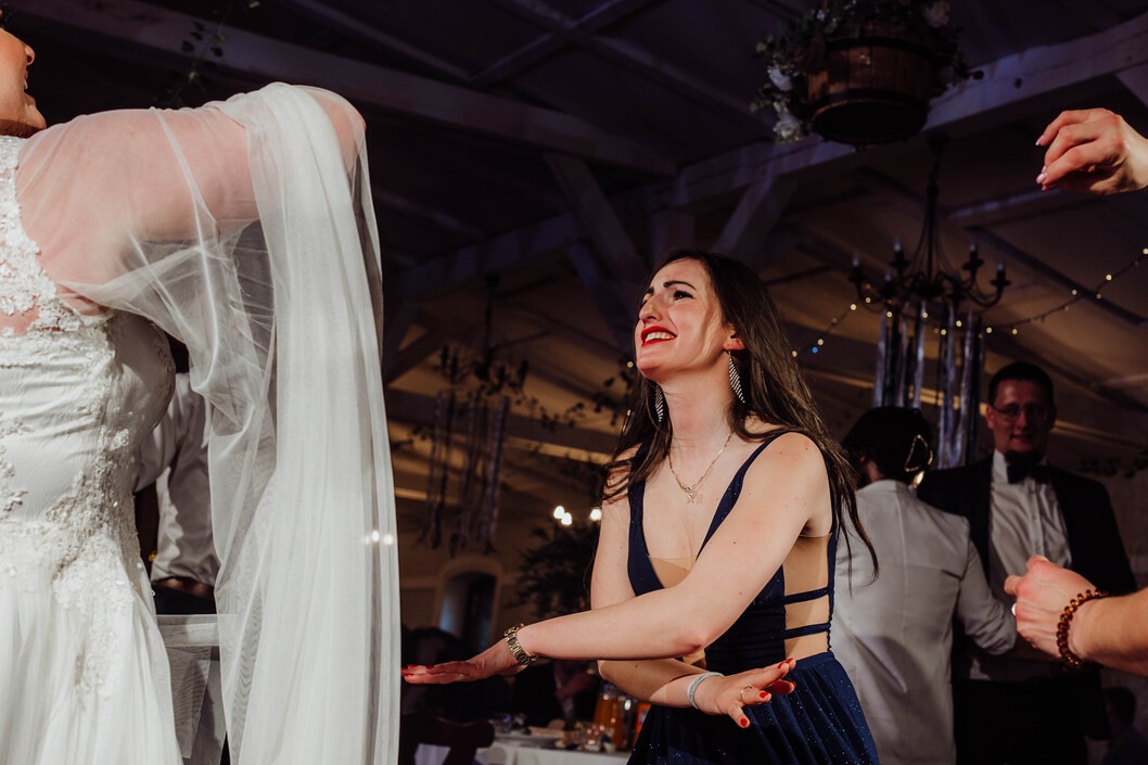 Panna młoda w białej sukni i gość w granatowej sukience tańczą podczas wesela, w tle widoczny mężczyzna w garniturze i dekoracje sali.