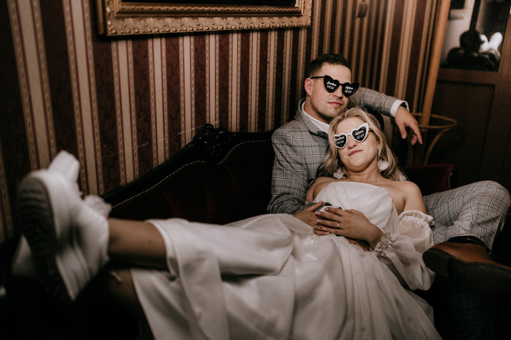 Para młoda w okularach w kształcie serc z datą ślubu, leżąca na bordowej sofie na tle ściany w paski, panna młoda ma założone białe sportowe buty.