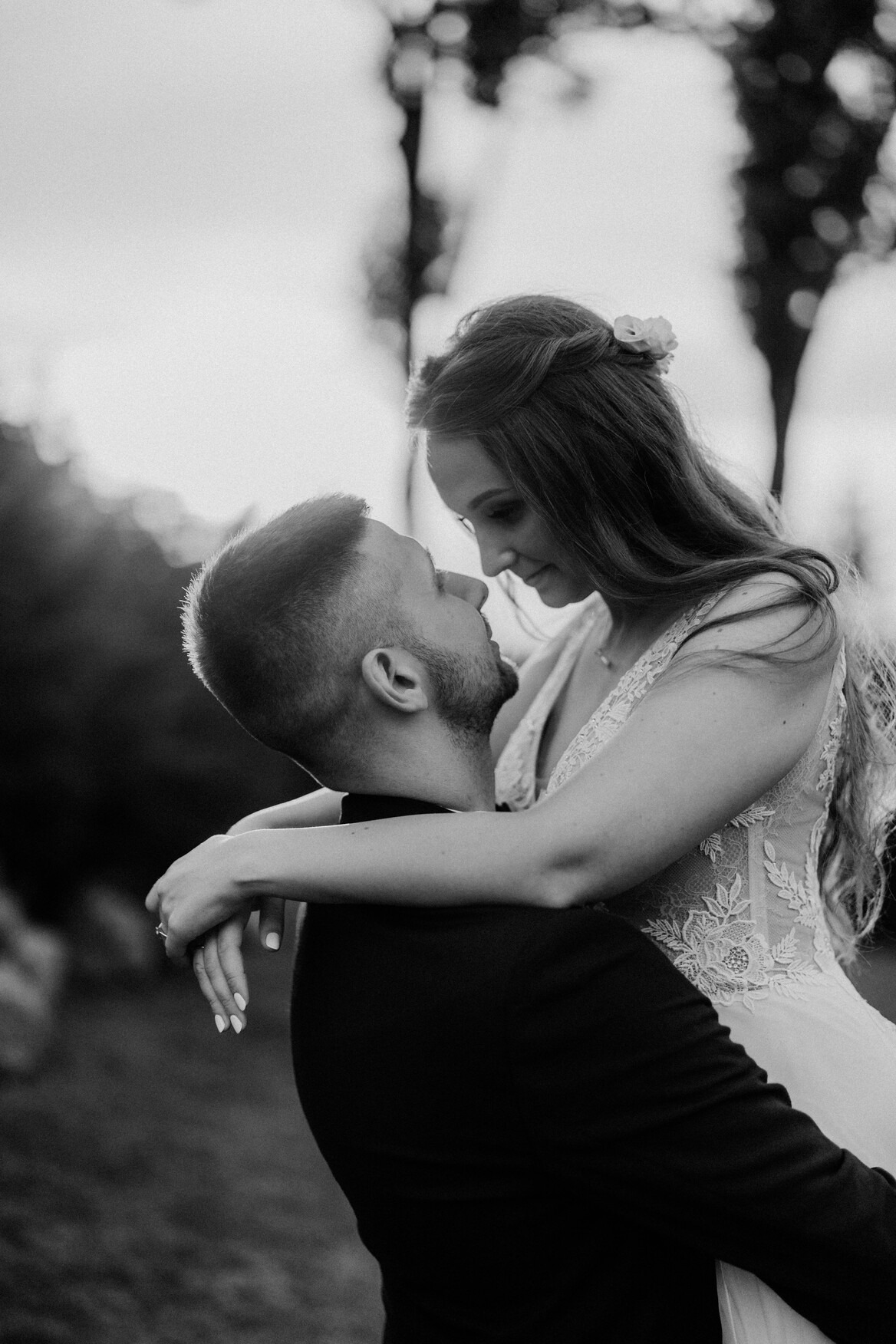 Czarno-białe zdjęcie: Panna młoda w koronkowej sukni z kwiatem we włosach, obejmująca pana młodego w czarnym smokingu, patrzą na siebie z bliska.
