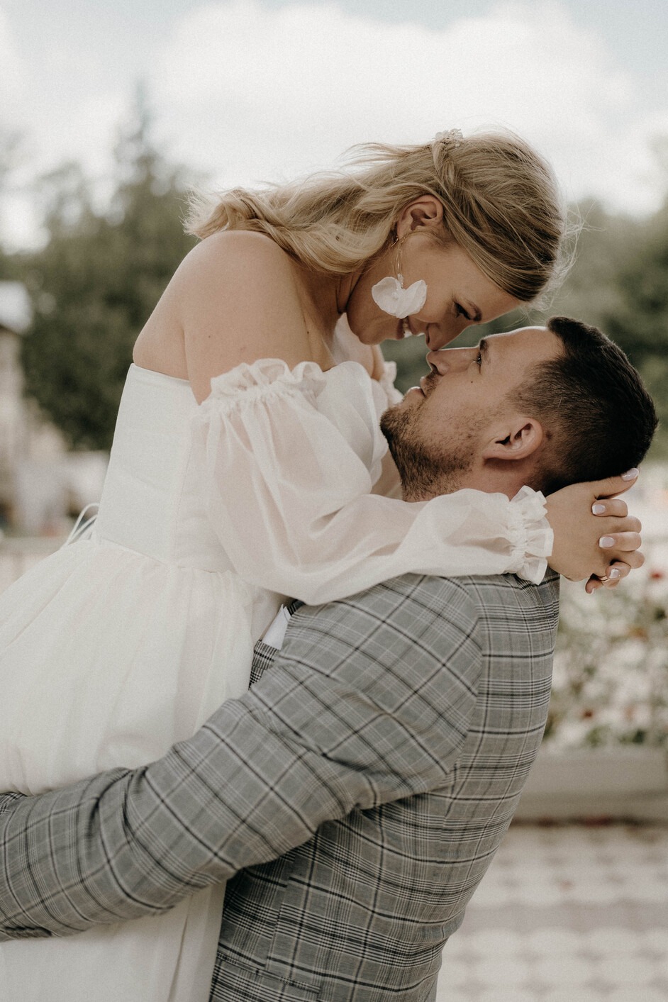 Panna młoda w białej sukni z bufiastymi rękawami i kolczykami w kształcie płatków obejmowana przez pana młodego w szarej kraciastej marynarce, zbliżający się do pocałunku na tle zieleni.