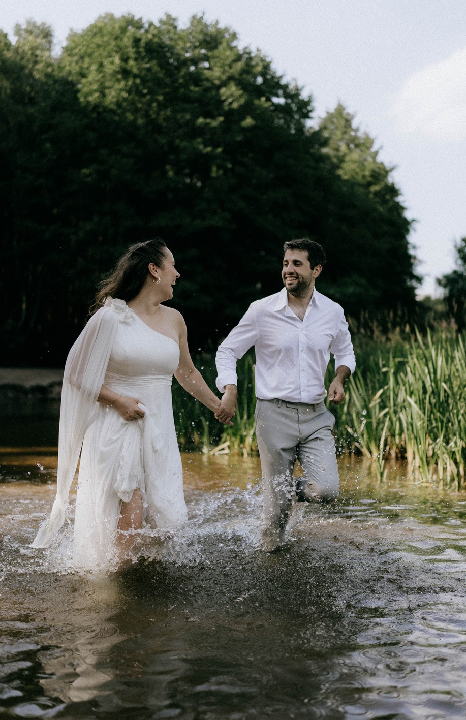 Para młoda w białych strojach biegnie trzymając się za ręce przez płytką wodę, tworząc rozbryzgi w promieniach słonecznych, z zielonym tłem drzew.