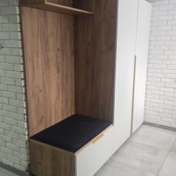 Adrian Jarząb - Nowoczesna zabudowa przedpokoju z drewnianymi panelami i białymi szafami, wnęką z czarną tapicerowaną ławką, otwartą półką i szufladą, na tle białej ceglanej ściany.