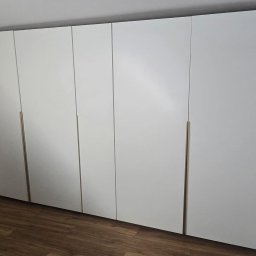 Adrian Jarząb - Biała, sześciodrzwiowa zabudowa meblowa z minimalistycznymi, pionowymi uchwytami w kolorze złotym, idealnie dopasowana do ściany.
