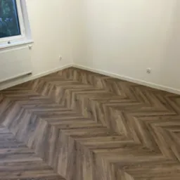 Ułożona podłoga z paneli imitujących drewno w jodełkę, widoczny fragment białego grzejnika pod oknem i białe listwy przypodłogowe.