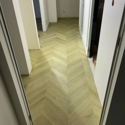 Fishka Floors Jacek Fiszka-Borzyszkowski - Jasna, nowa podłoga ułożona we wzór jodełki w korytarzu, widok zza otwartych drzwi, częściowo widoczne inne pomieszczenia w tle.
