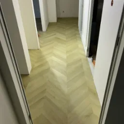 Jasna, nowa podłoga ułożona we wzór jodełki w korytarzu, widok zza otwartych drzwi, częściowo widoczne inne pomieszczenia w tle.