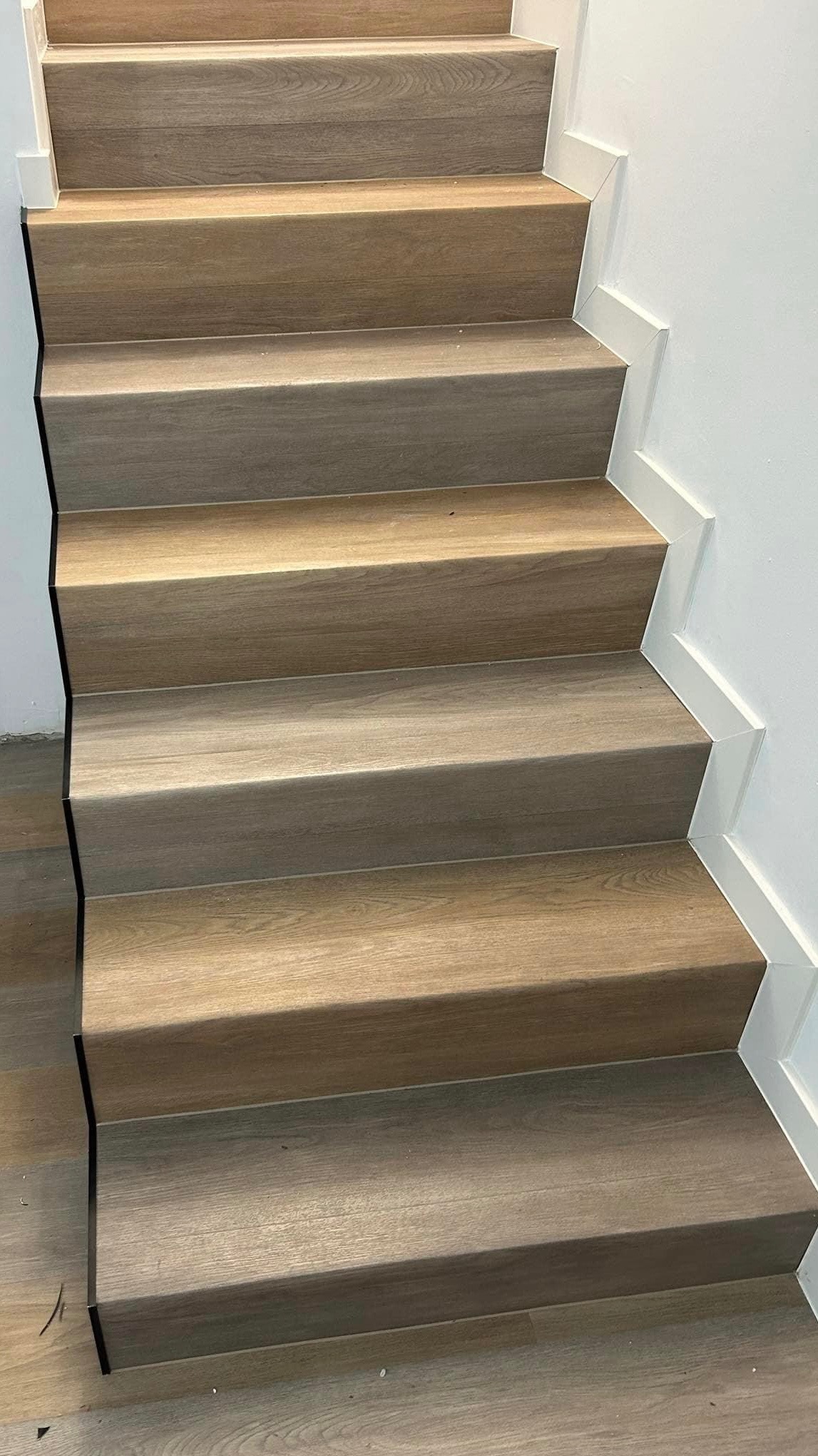 Schody wykończone panelami podłogowymi w odcieniach beżu i brązu, z białymi listwami przypodłogowymi po prawej stronie, widok z dołu.