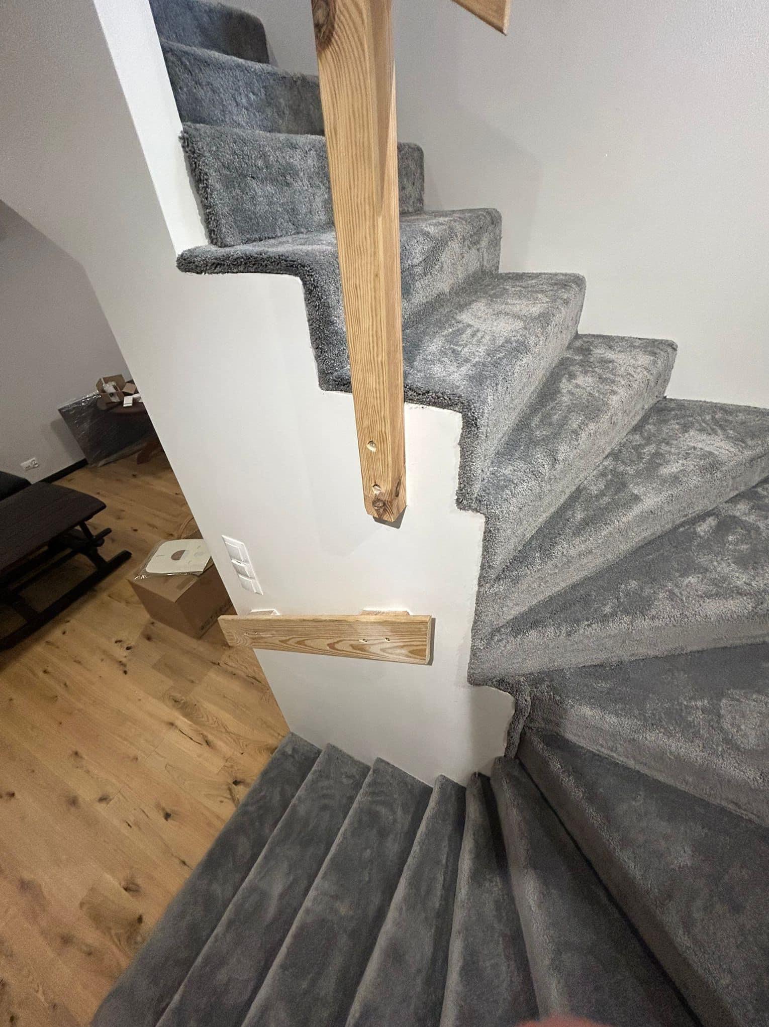 Spiralne schody z szarego dywanu, drewniana balustrada, widoczne wykończenie podłogi drewnianej w tle.