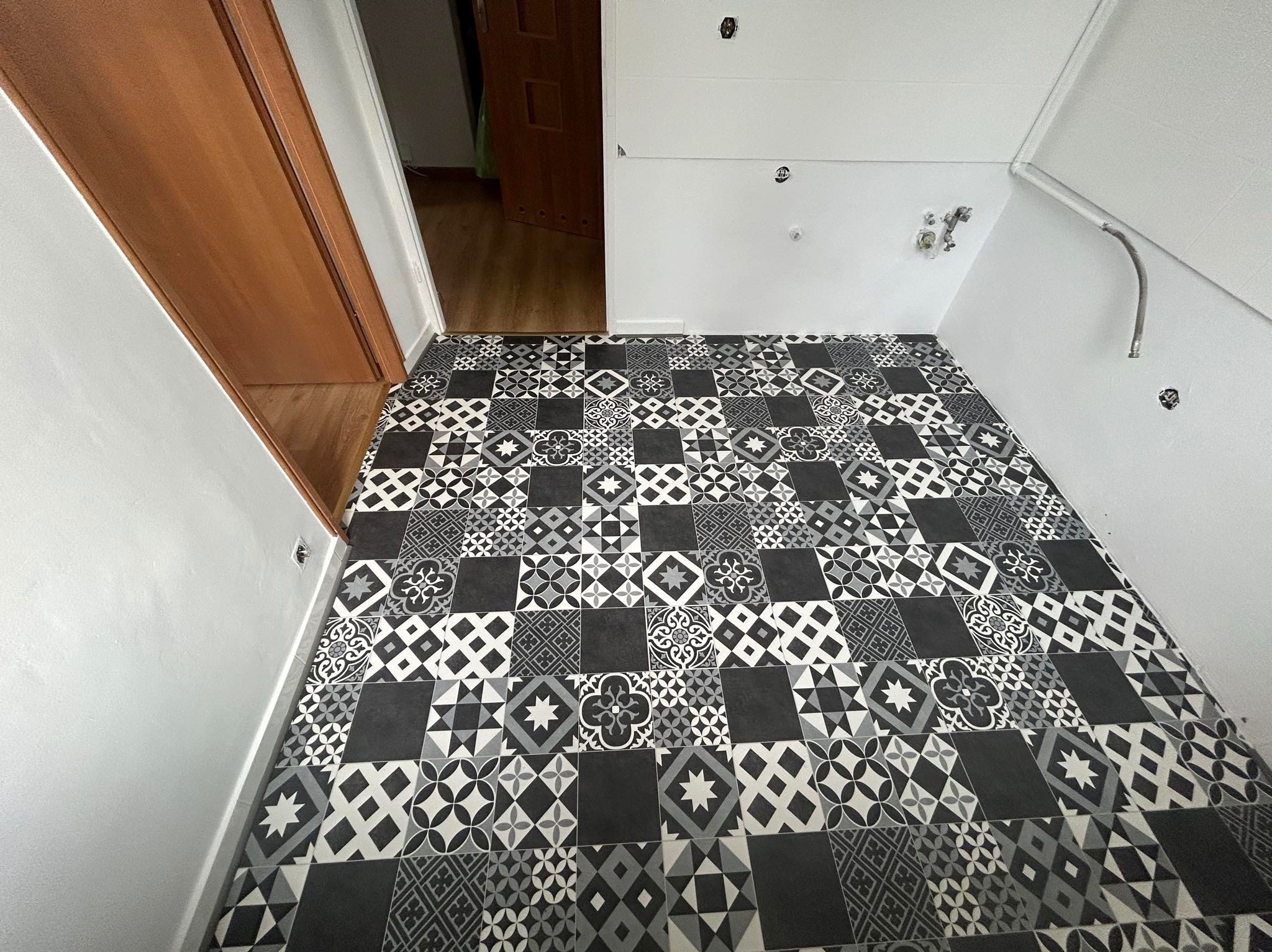 Ukończona podłoga w małym pomieszczeniu wyłożona czarno-białymi płytkami o różnych wzorach geometrycznych i kwiatowych, widoczna część ściany z instalacją hydrauliczną, drzwi z drewnianą ościeżnicą...