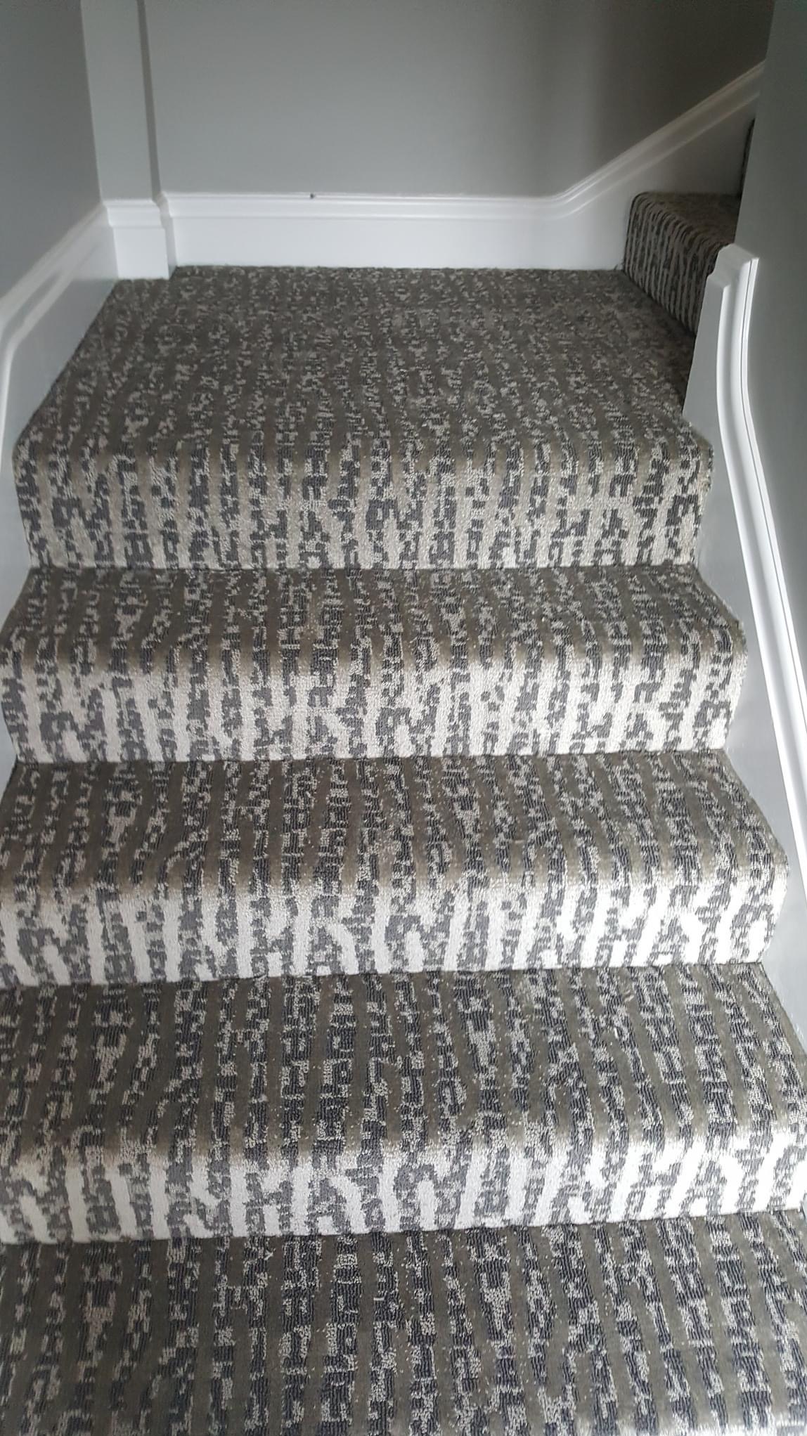 Schody pokryte wzorzystą wykładziną dywanową w odcieniach szarości i beżu, widok z dołu, białe listwy przypodłogowe