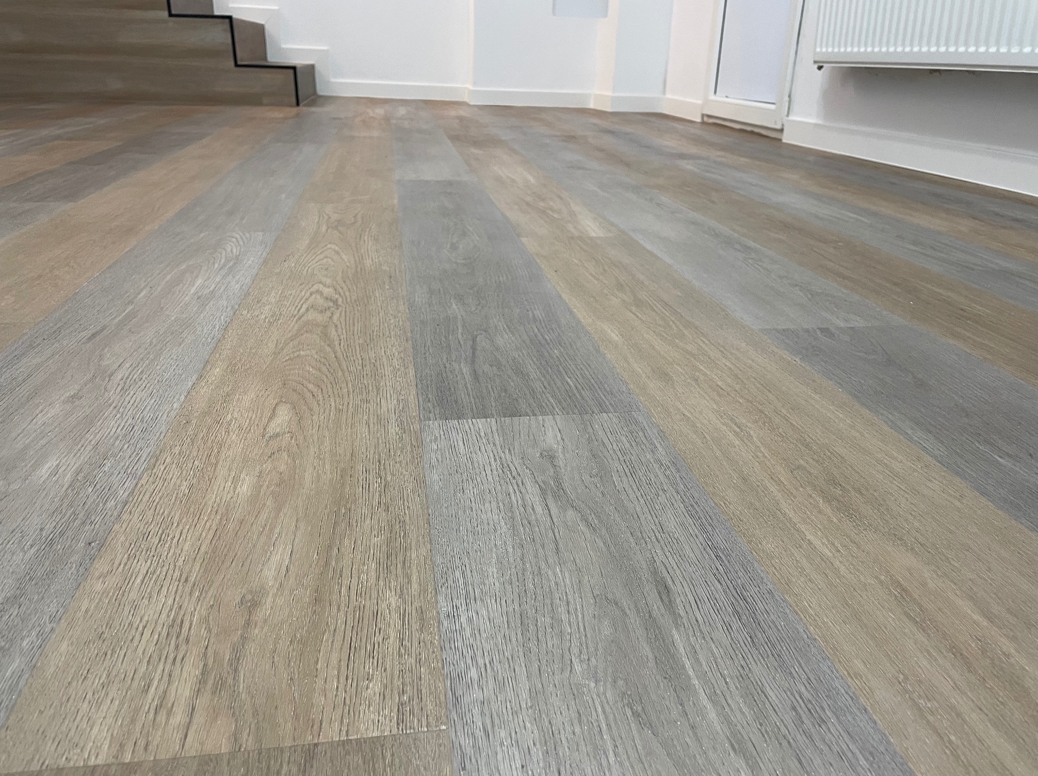 Ułożone panele podłogowe w odcieniach szarości i beżu, widok z niskiej perspektywy, z fragmentem schodów i grzejnikiem w tle.