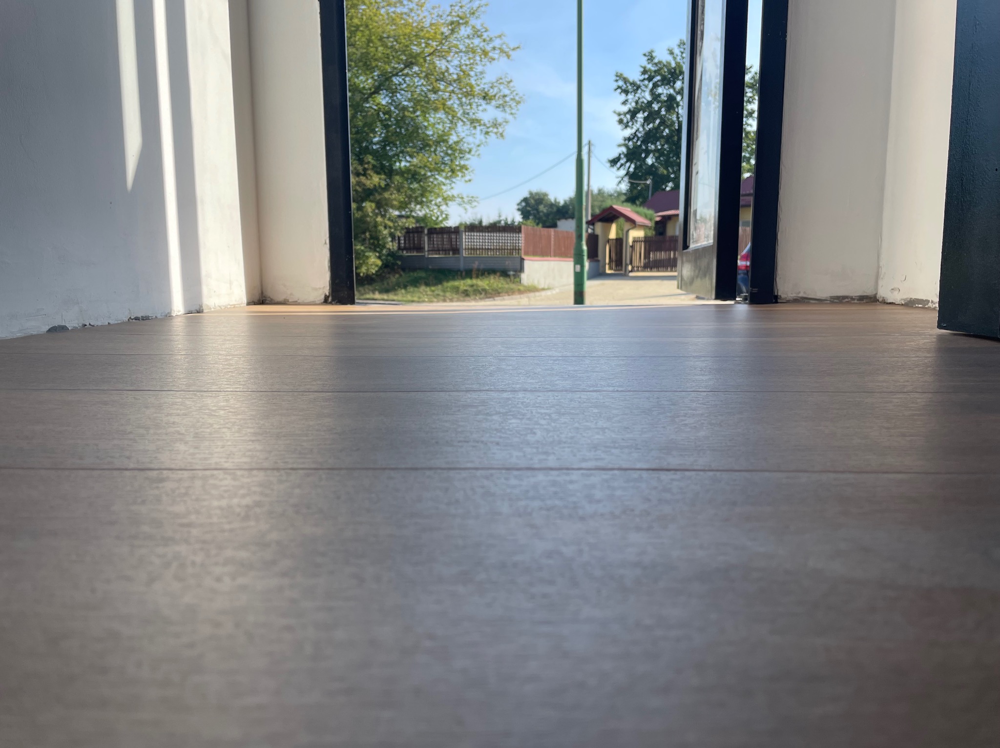 Nowa, ciemna podłoga z paneli, widok z niskiej perspektywy na otwarte drzwi i fragment podwórza z zielenią.