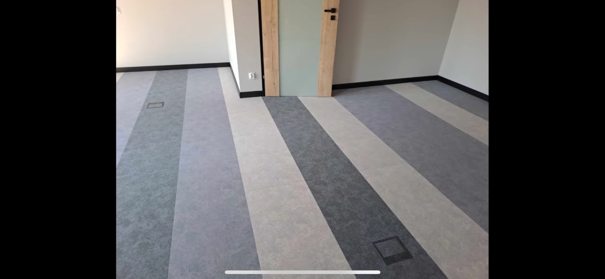 Wnętrze z podłogą w pasy z paneli w odcieniach szarości, z widocznymi kratkami wentylacyjnymi w podłodze i drzwiami w tle.